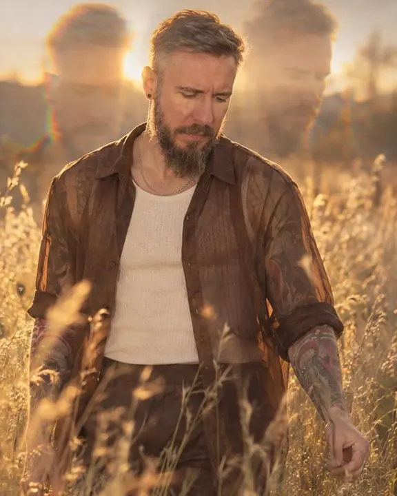 A man in a sheer brown organza shirt walks slowly through tall grass, his head and shoulders repeated above