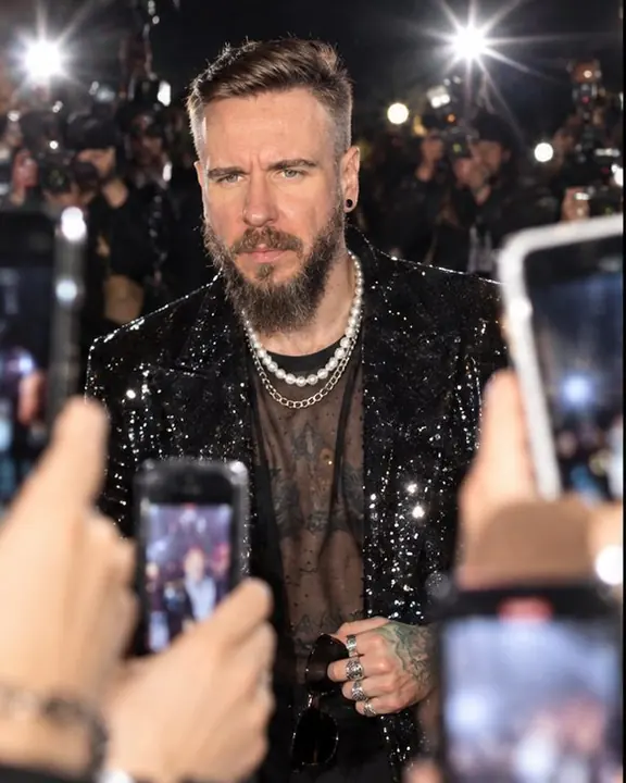 A bearded man in a black sequin coat walks into a storm of flashes, face sharp, five phones in the foreground