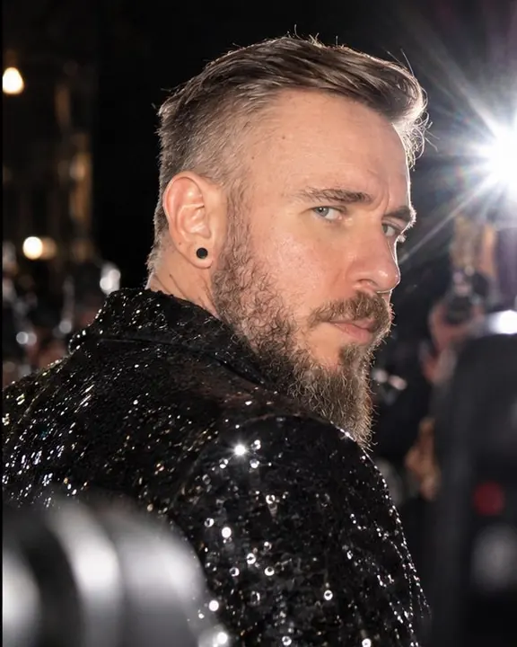Over-the-shoulder close portrait of a bearded man glancing back at cameras, flashes reflecting in sequins