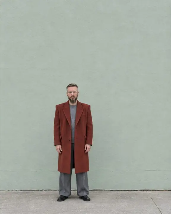 A bearded man in a rust-red coat stands small in the lower third of a large pale mint wall, arms at his sides