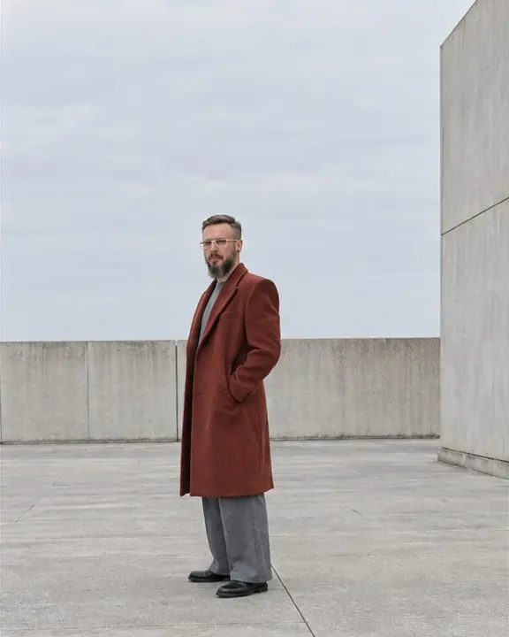 A bearded man at the far edge of an empty concrete plaza, turned three-quarters, one hand in his coat pocket