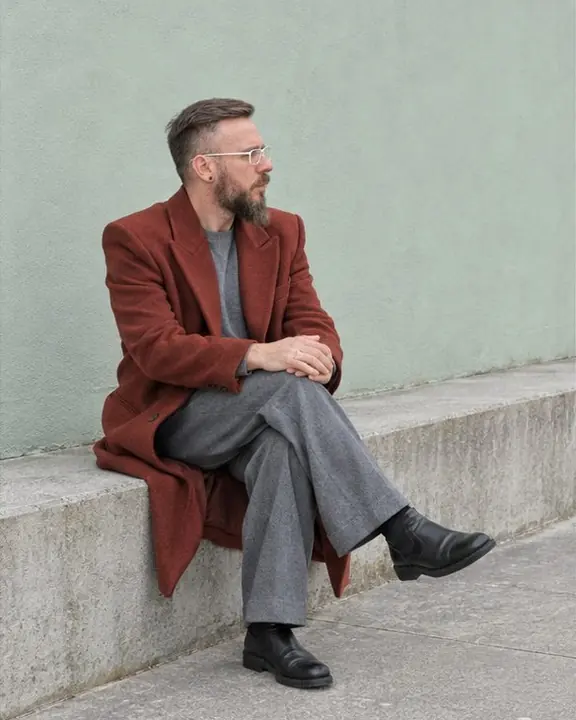 A man in a long red coat sits on a low concrete ledge with legs crossed, looking away along a pastel wall