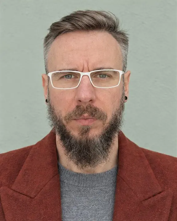 Head-and-shoulders portrait of a bearded man in white rectangular glasses, calm frontal gaze, squared shoulders below