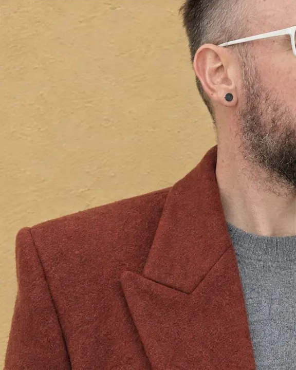 Close crop of a squared rust-red coat shoulder seam and collar against a pale wall, the side of a face at the edge