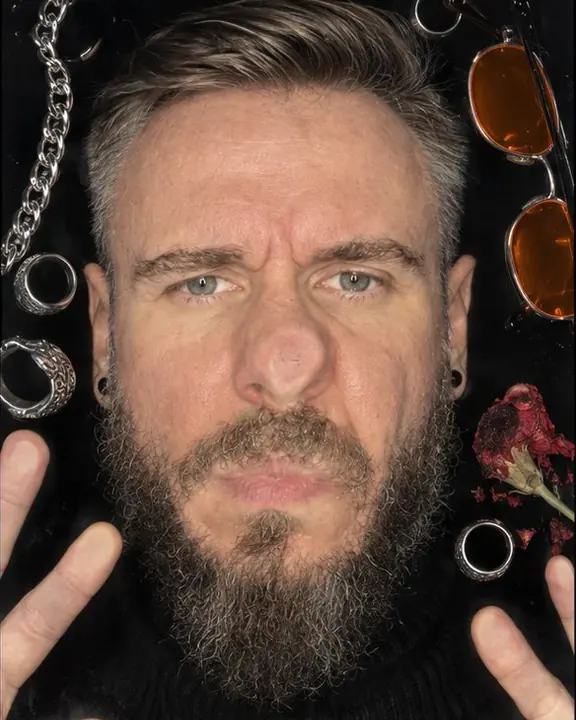 A bearded man's face pressed flat against scanner glass, nose and cheek flattened, silver rings and chains around