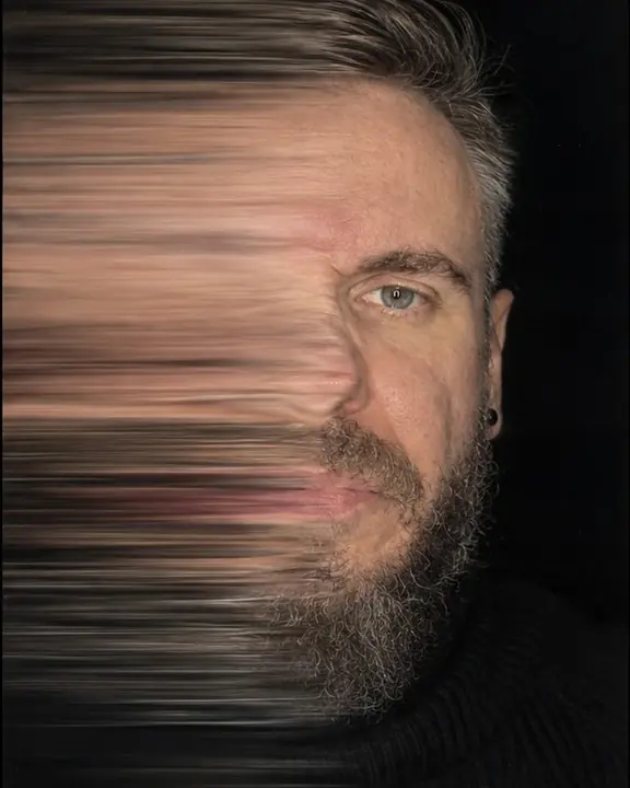 A bearded face pulling back during a scan, one side stretched into streaks, one eye sharp against black