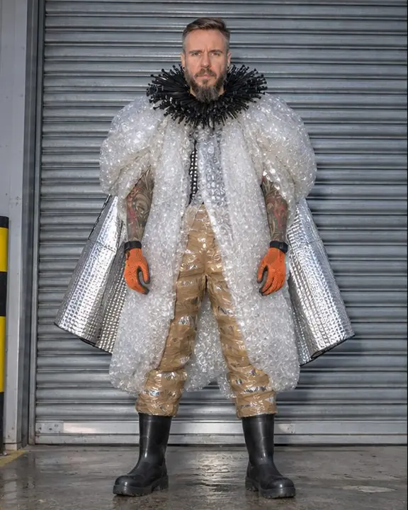 A bearded man in a huge bubble-wrap cocoon coat stands frontally before a corrugated metal shutter