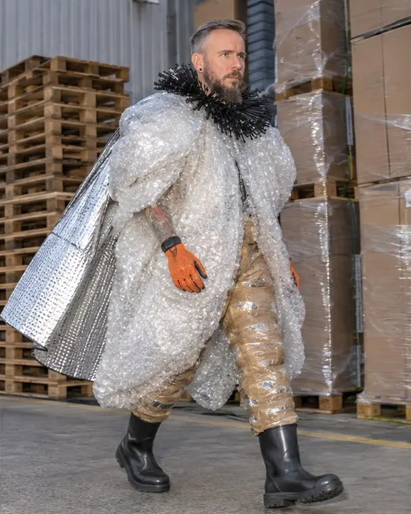 A man walks past stacked pallets in three-quarter view, a silver car-sunshade cape catching the flash
