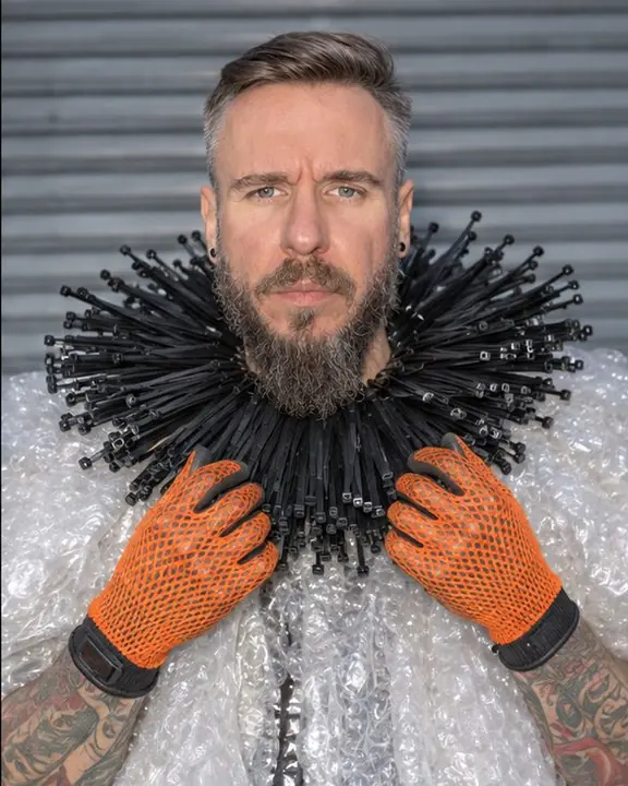 Waist-up of a bearded man framed by a stiff ruff of black cable ties, looking straight into the lens