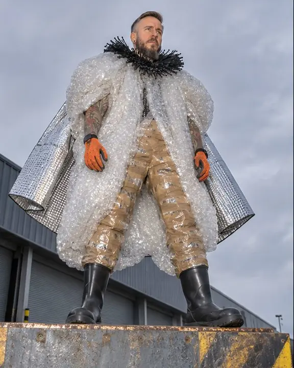 Low-angle full-length of a man in bubble wrap and tape pants on a loading dock edge, gray sky behind