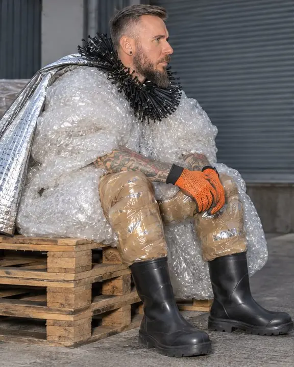 Side profile of a man seated on a pallet, shiny packing-tape pants, orange mesh gloves resting on his knees