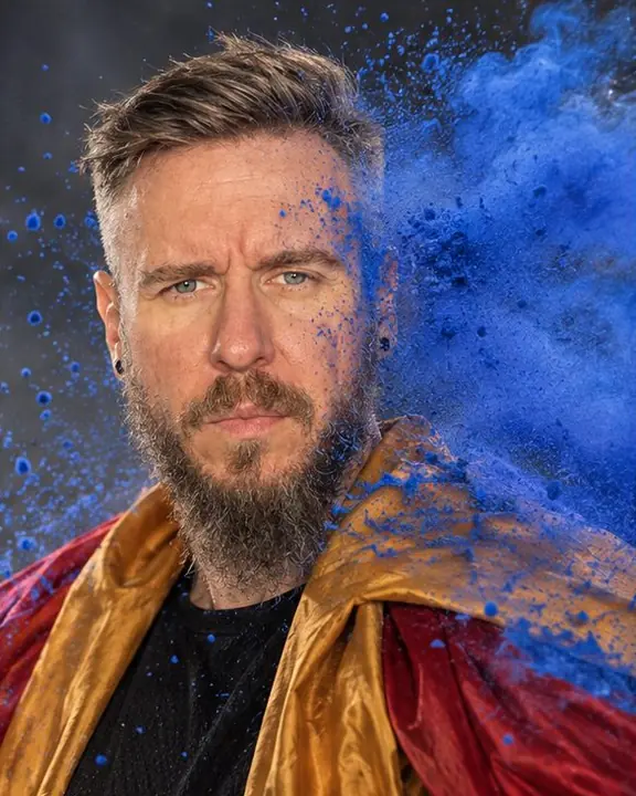 Waist-up of a man with eyes open as a cloud of ultramarine powder explodes past his face, frozen by flash