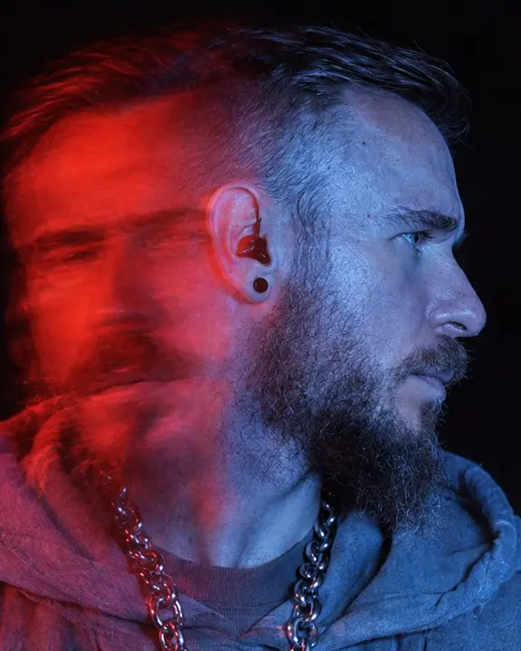 Close profile of a bearded man in blue light, a red smeared frontal version of his face overlapping it