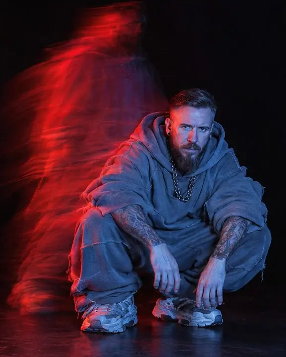 A bearded man crouching sharp in blue while a red ghost of him standing up lingers above, black studio