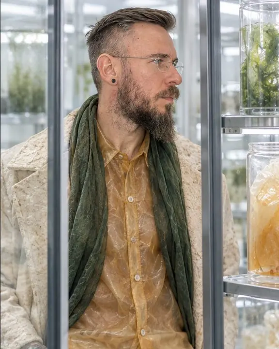 Seen through a glass incubator wall, a man in a translucent amber cellulose shirt glows in cool, even light