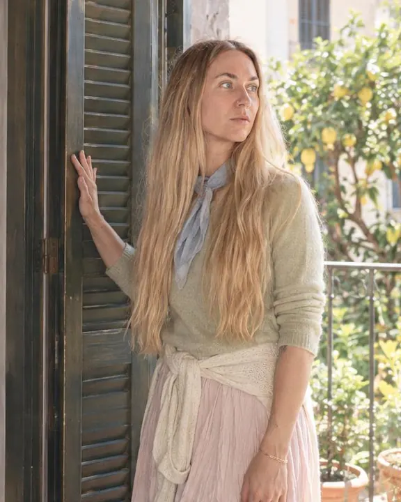 Three-quarter view of a woman with long blond hair by tall open shutters, hand on one, head turned toward a lemon tree