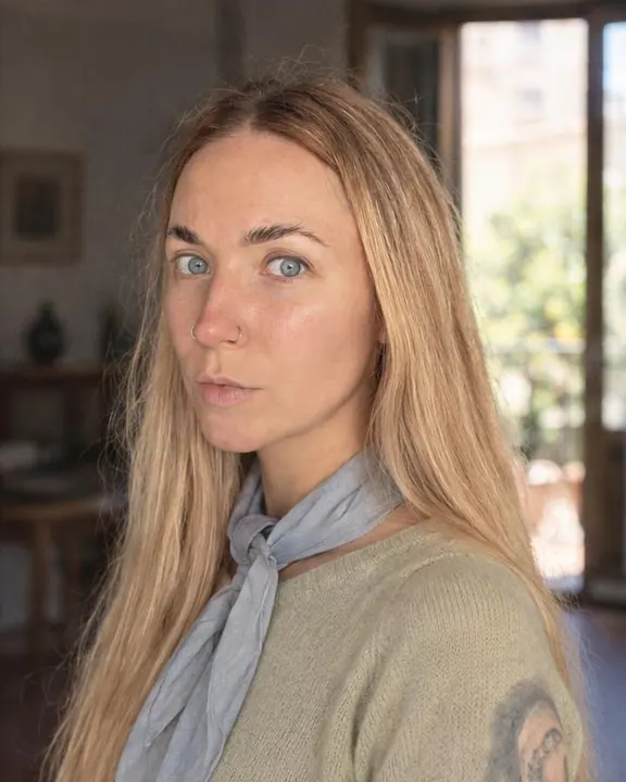 Waist-up in a dim white room, a woman with long blond hair looks into the lens, a powder-blue silk scarf loose at her neck