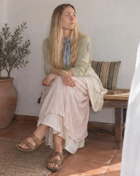 A woman with long blond hair sits on a low wooden bench against a white wall, ankles crossed in tan strap sandals