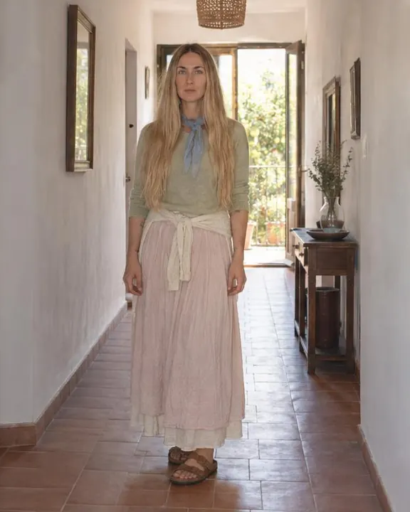 A woman with long blond hair stands small in a long white corridor, the bright doors behind outlining her pink skirt