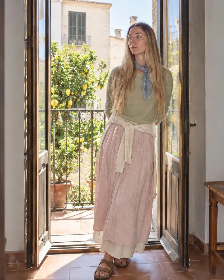 A woman with long blond hair leans in an open balcony doorway, backlight rimming her pistachio knit, face soft in fill flash