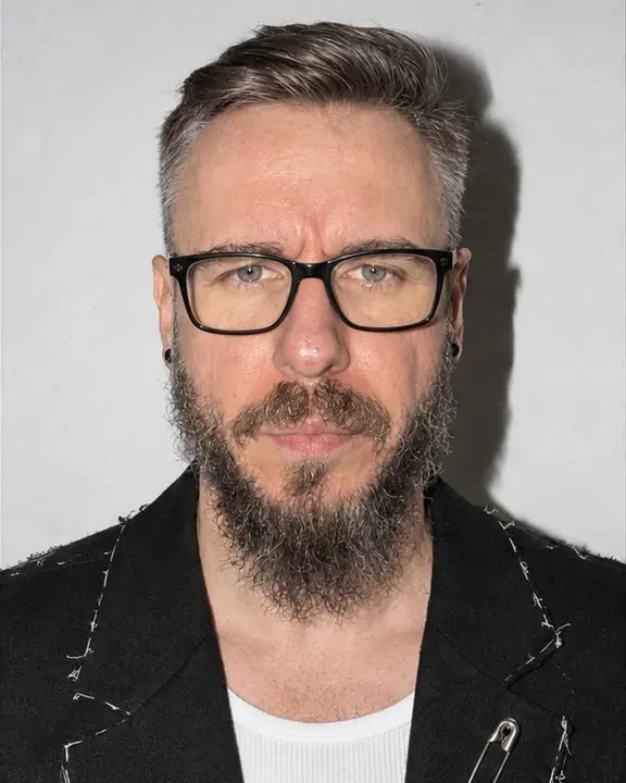 Tight portrait of a bearded man in black glasses against a white wall, eyes to the lens, a hard flash shadow behind