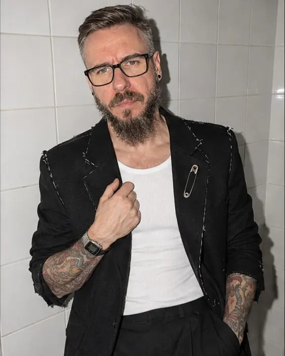 A man in a raw-edged black blazer leans on white bathroom tiles, one hand at the lapel, lit by a bare flash