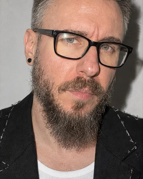 A bearded man mid-turn toward the lens, cropped at forehead and chest, a small flash glint at the edge of his glasses