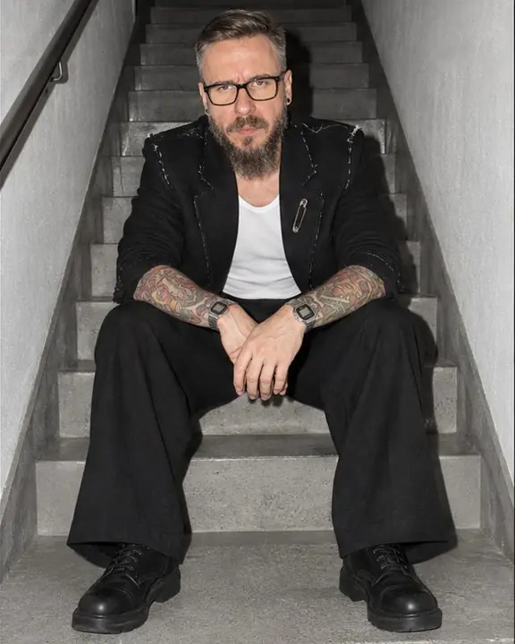 A man in black tailoring sits on stairwell steps, elbows on knees, the flash falling off into darkness above
