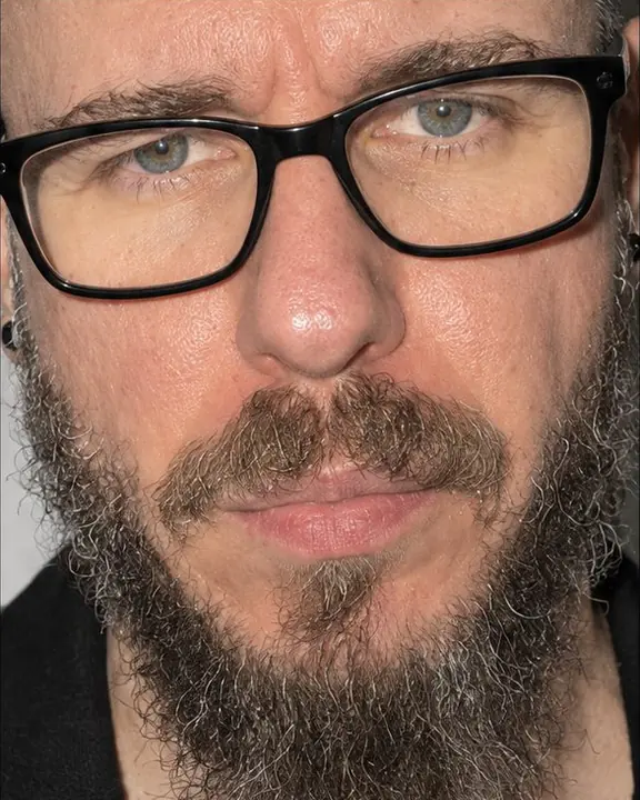 Extreme close-up from eyebrows to chin of a bearded man in thick black frames, skin and hair texture in hard flash