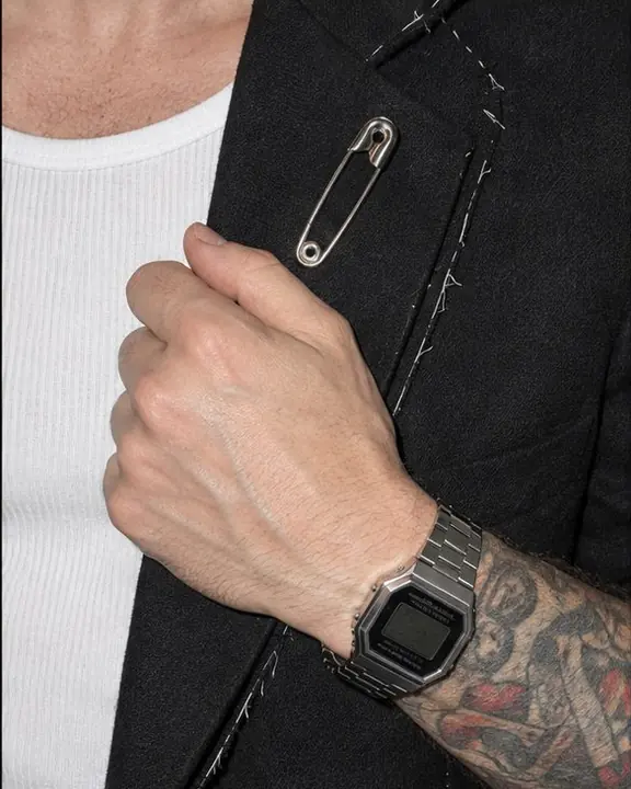 Hard-flash close-up of a hand at a raw blazer lapel with a safety-pin brooch and a steel digital watch