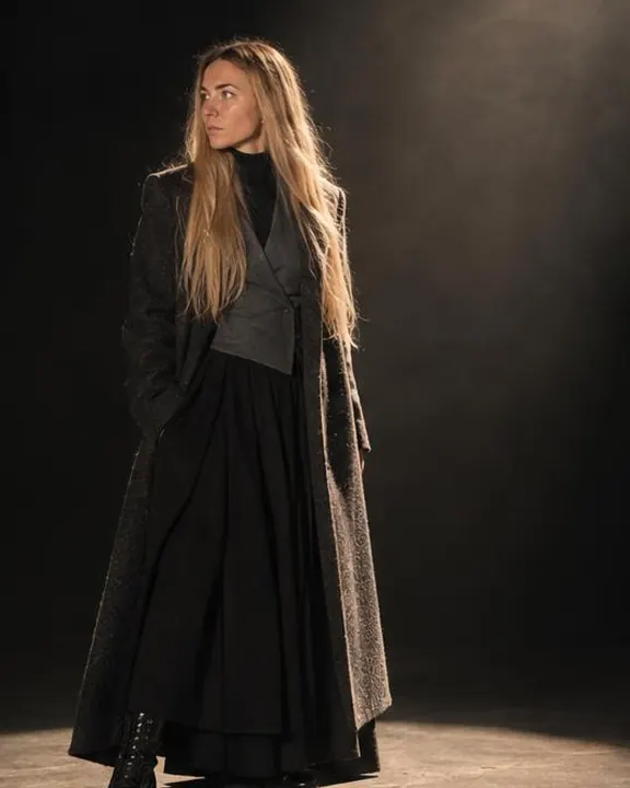 A woman with long blond hair in a charcoal coat steps one foot into a spotlit oval, the coat hem lit, the rest in darkness