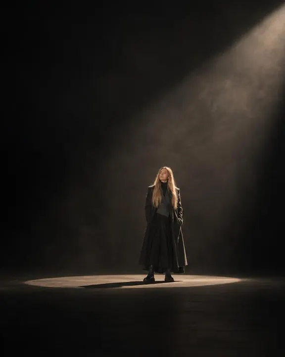 Wide view of a woman with long blond hair small inside a lit floor oval, a visible cone of light falling through studio haze