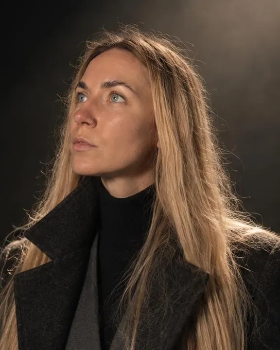 A woman with long blond hair turns her face up into a hard spotlight beam, a clean shadow under the cheekbone, collar lit