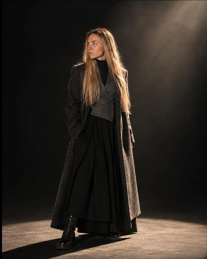 A woman with long blond hair in a charcoal coat steps one foot into a spotlit oval, the coat hem lit, the rest in darkness