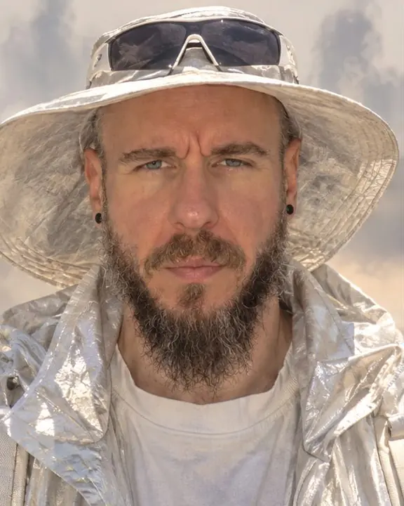 Head and shoulders of a bearded man in a wide sun hat, face readable while the background boils with haze
