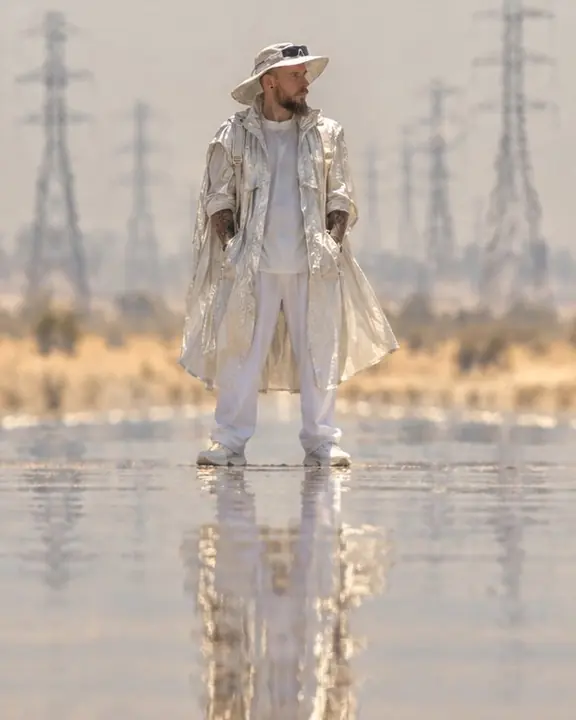 A man stands on a desert road with power pylons compressed behind him, doubled below by a mirage