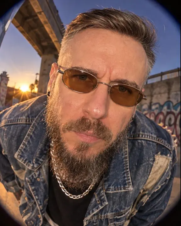 A bearded man leans into a fisheye lens inches away, face in the center, curved buildings bending behind him