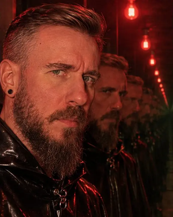 Head and shoulders of a bearded man in red light, a tunnel of his reflected faces receding behind him