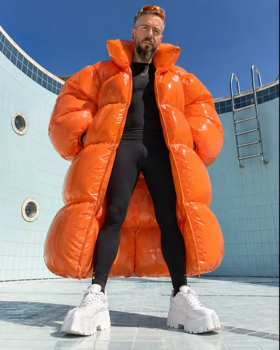A bearded man in a huge glossy orange inflatable coat stands in the deep end of an empty pool against pale tiles