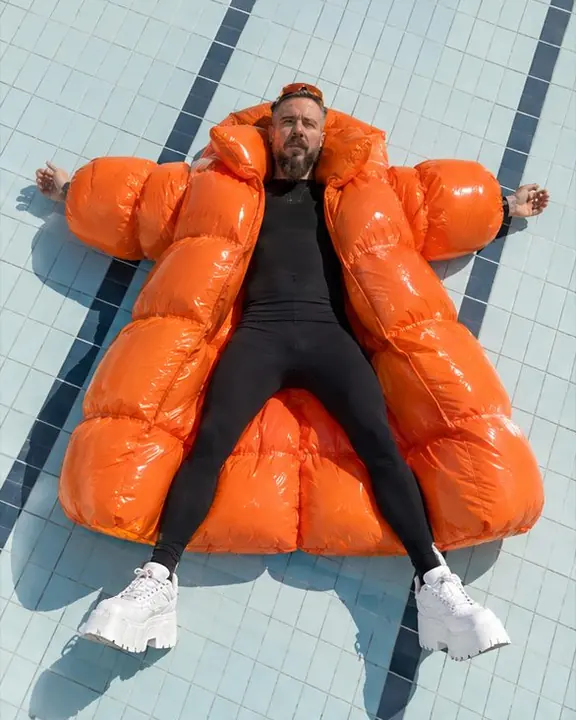 High-angle view of a man lying on a pale aqua pool floor in an inflated orange coat, like a giant orange shape