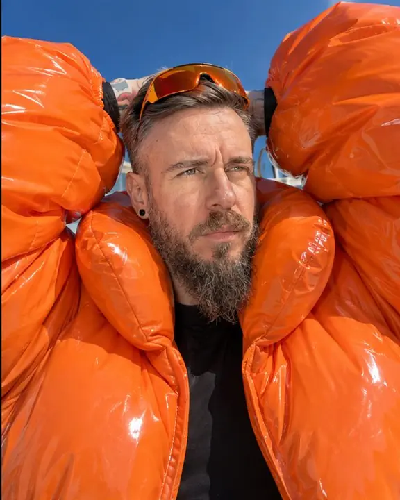 A bearded man raises his arms so huge puffed orange sleeves frame his face under a cobalt sky