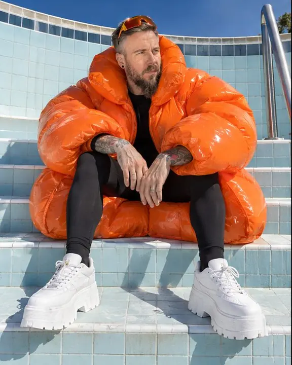 A man sits on empty pool steps, the glossy orange coat bulging around him, a long shadow on the tiles