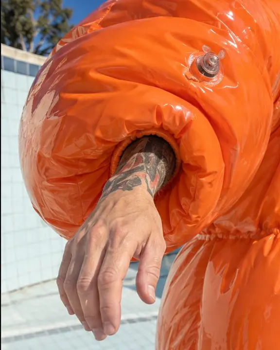 A glossy orange PVC sleeve reflecting sky and pool tiles, a hand emerging from it in hard sun