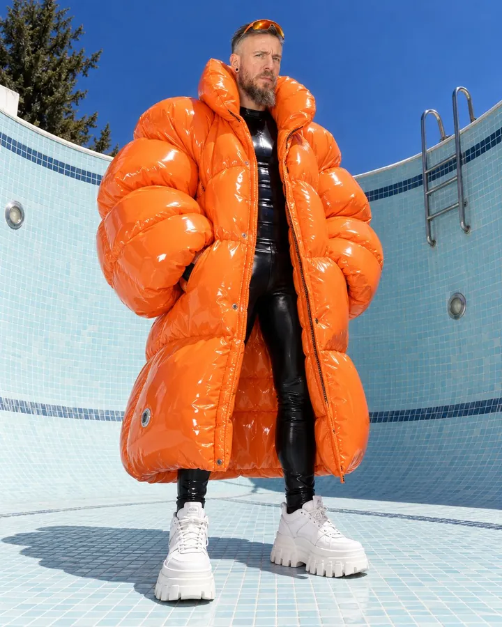 A bearded man in a huge glossy orange inflatable coat stands in the deep end of an empty pool against pale tiles