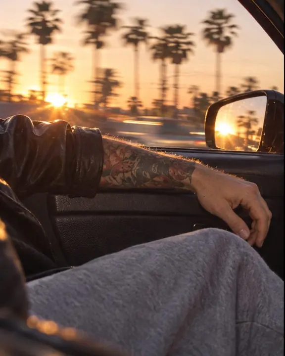 A man's arm in black leather resting on a car door, chunky slides on the dashboard, palms streaking past outside