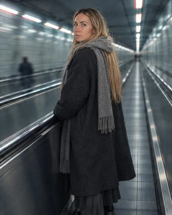 From behind and to one side, a woman with long blond hair in a charcoal coat and gray scarf, tubes drawn into cyan lines