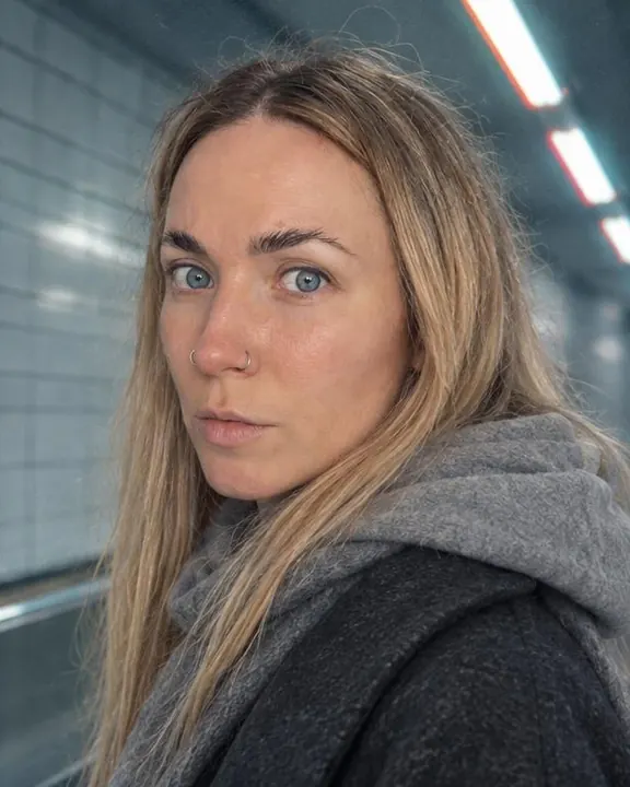 Side view of a woman with long blond hair facing the lens, a gray hood bunched at her collar, a red glow on the tube behind
