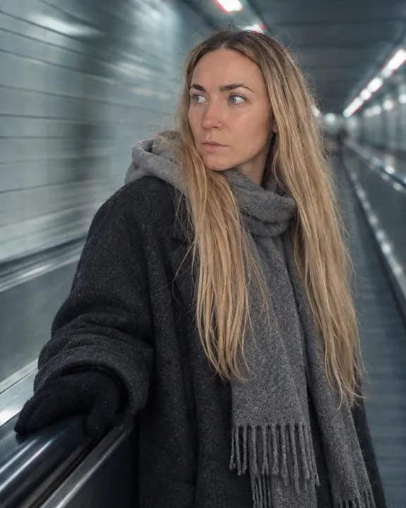 Waist-up, a woman with long blond hair rests a hand on the rail and looks down the cyan tunnel past the camera, collar up