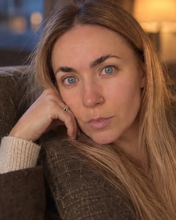 A woman with long blond hair rests her head on the sofa back, silver ring by her jaw in lamp light, the other cheek blue