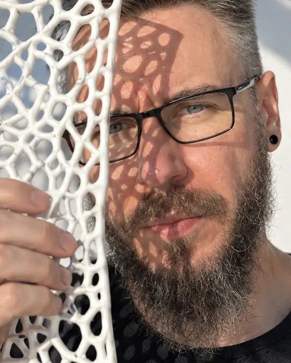 A bearded man holds a printed lattice panel near his face so its sharp shadow crosses his cheek